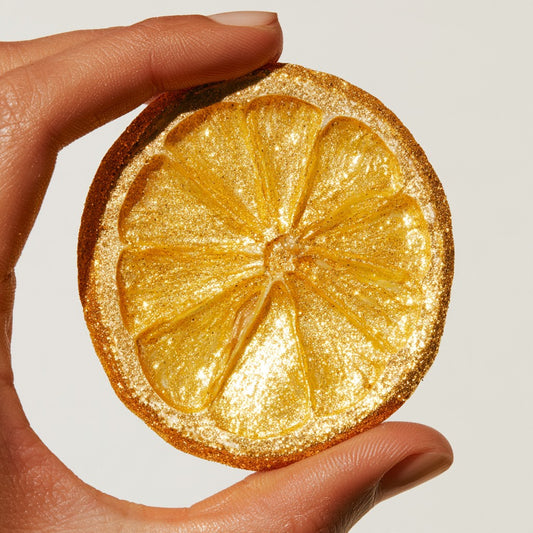 Main tenant une tranche de citron jaune séchée pailletée d'or sur fond blanc | The Originea