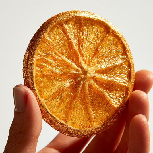 Main tenant une tranche d'orange séchée pailletée d'or sur fond blanc | The Originea